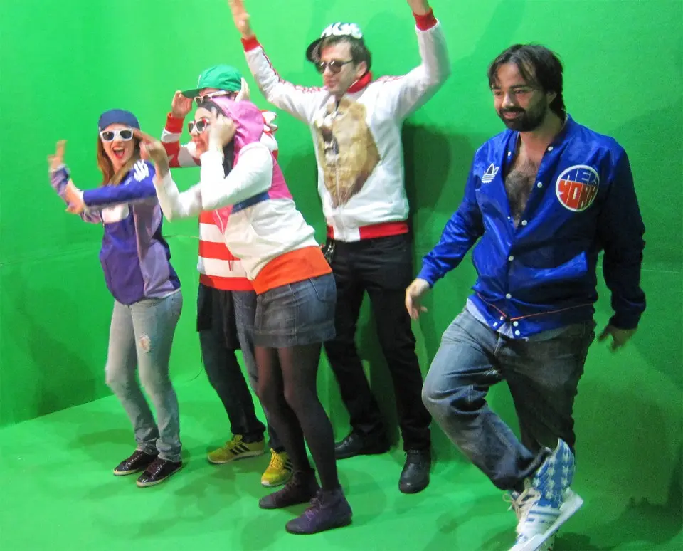 Five people dancing in front of a green screen, with colorful clothes and raised arms