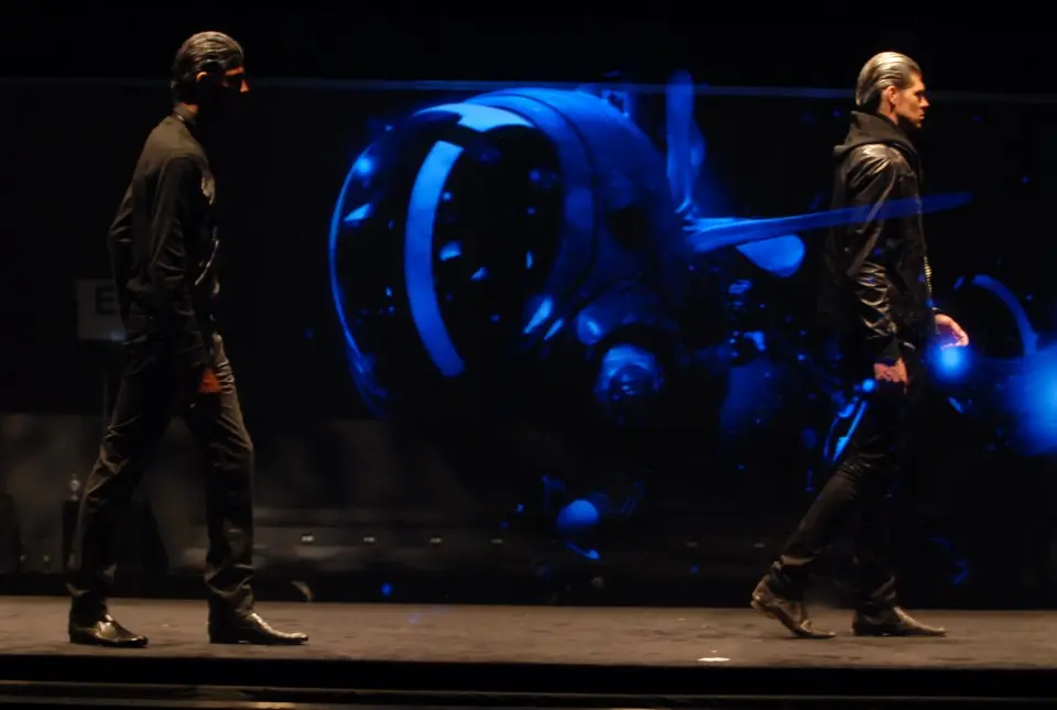 Two models walking in opposite directions on the runway past a blue holographic shape