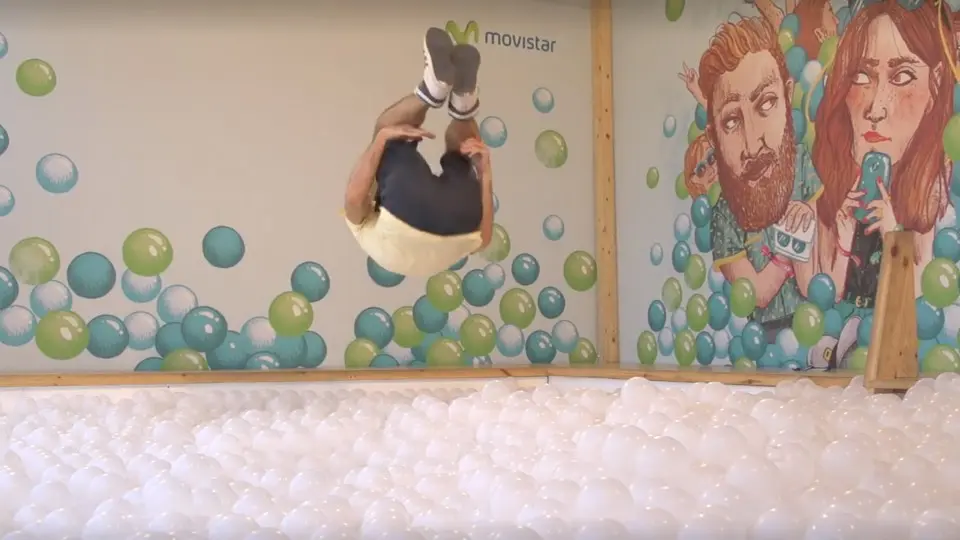 Person jumping head first into a ball pit in front of a mural of two illustrated faces