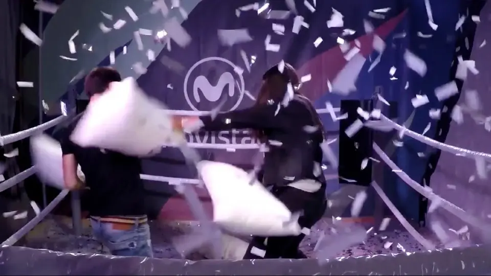 Two people having a pillow fight in a boxing ring while feathers and confetti fly through the air