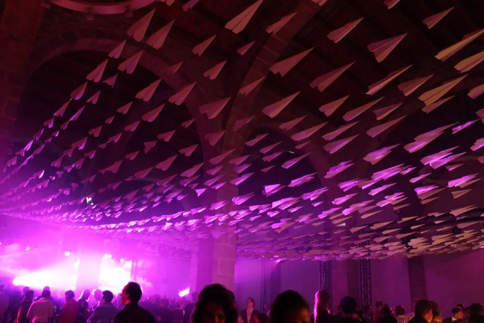 Dense layer of paper planes covering the ceiling above partygoers in purple light